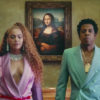 Still from Apeshit featuring Beyoncé in a purple suit next to Jay-Z in a turquoise suit, both looking forward with the Mona Lisa behind them