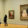 Three viewers with their back turned towards the photograph, looking at three portrait paintings in gilded frames at the Met