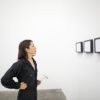 A lone, young woman in black coveralls holds a hammer and looks at a museum wall featuring three framed images