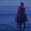 Woman in red sits atop a horse riding out of the blue ocean